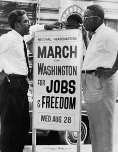 Bayard Rustin (left) and Cleveland Robinson (right), stand next to a sign identifying the National Headquarters for the March
