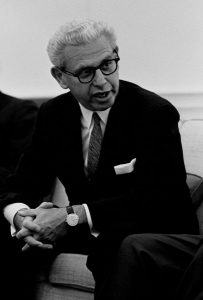 Arthur Goldberg sits on a couch with his hands clasped before him. He wears a suit, glasses and a watch; his hair is grey. He is speaking. 