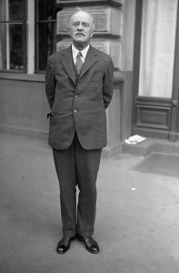 Oswald Parnson Villard stands in front of a building. His hands are clasped behind his back. He wears a suit.
