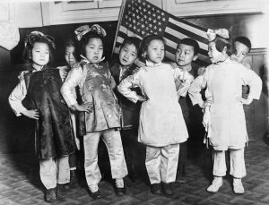 Chinese children in native costume stand before an American flag. 