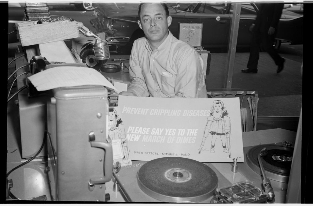 Man with sign raising money for the March of Dimes Foundation. He is sitting at the console of a radio station and appears to be the D J. 